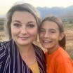 A woman in a striped, black blouse stands next to a young girl in an orange t-shirt in front of a mountain range.