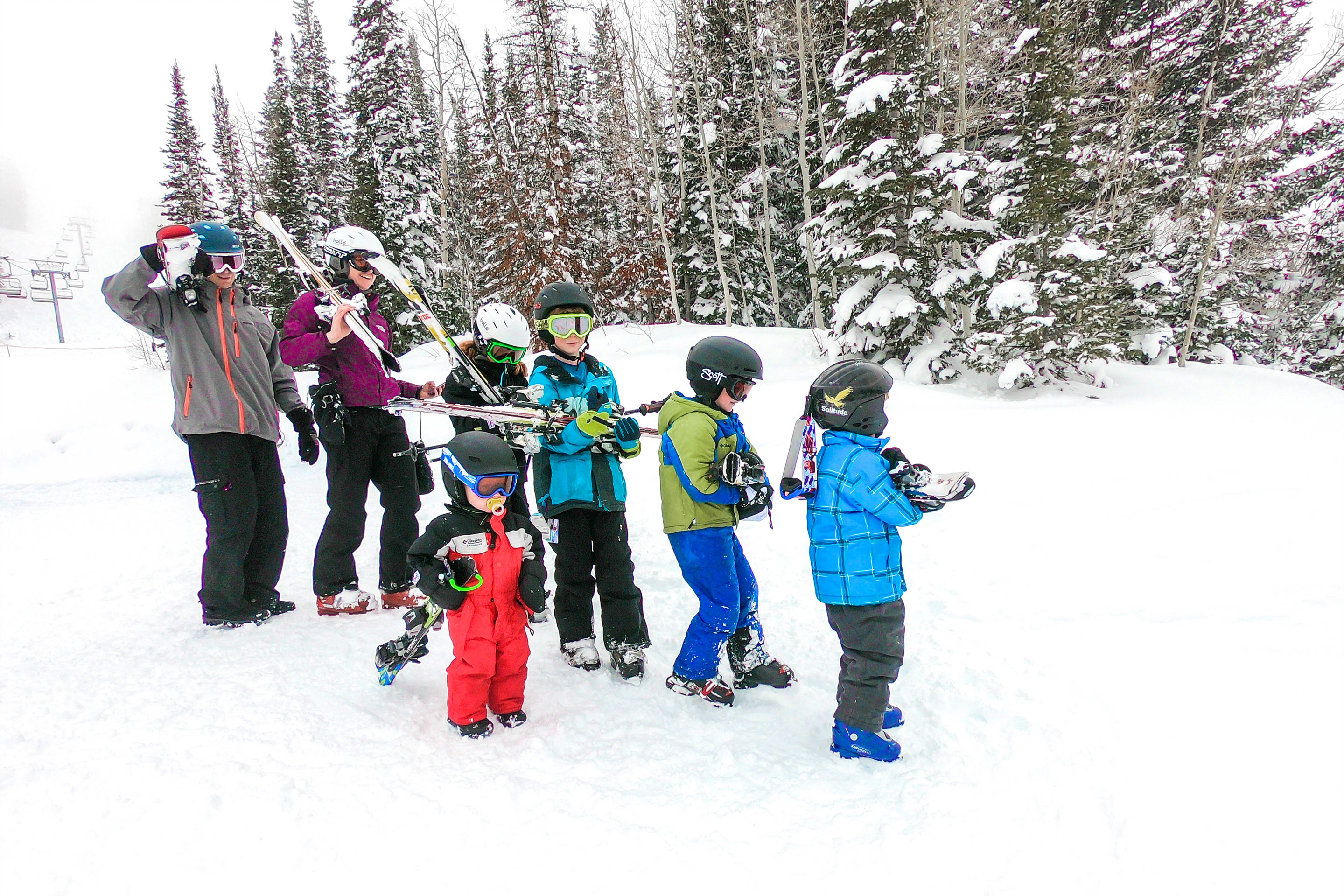 Two adults and five children wear winter apparel, ski helmets and goggle while holding a pair of ski's over their shoulders on a snowy slope.
