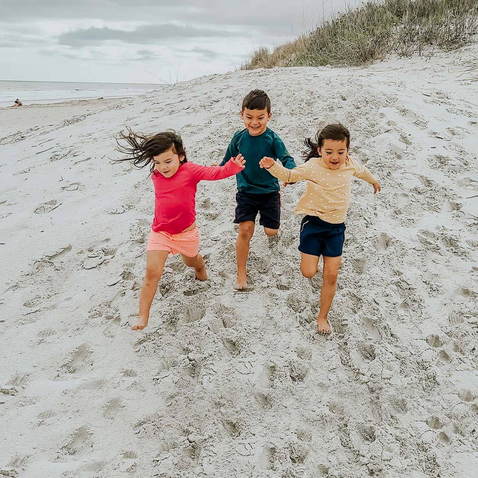 Three toddlers in multi-colored rash guards and board shorts run on a sandy beach.