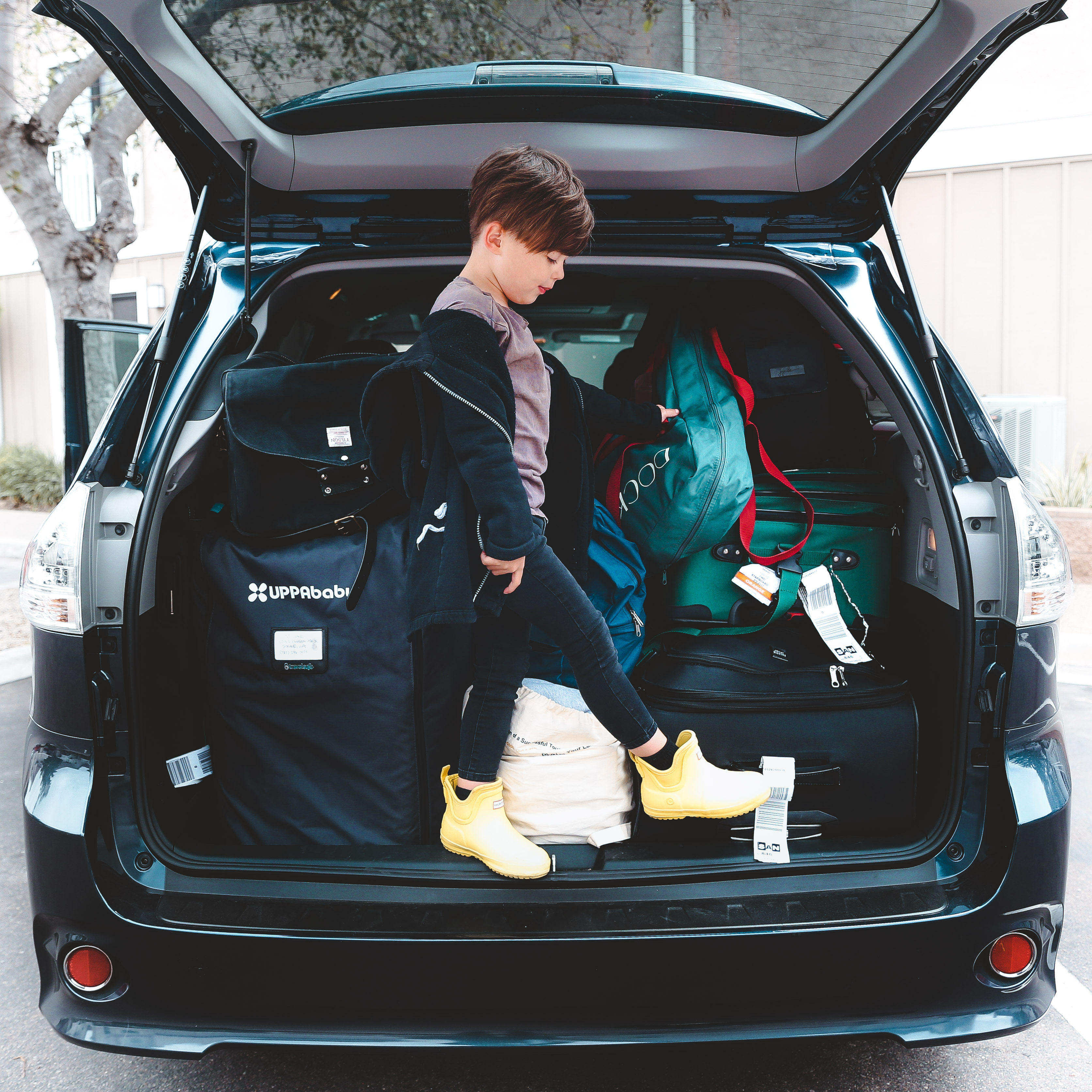Featured contributor, Mia St. Clair's son, Grey, sits on his white wheeled suitcase in an airport terminal wearing a black shirt and yellow rain booties.