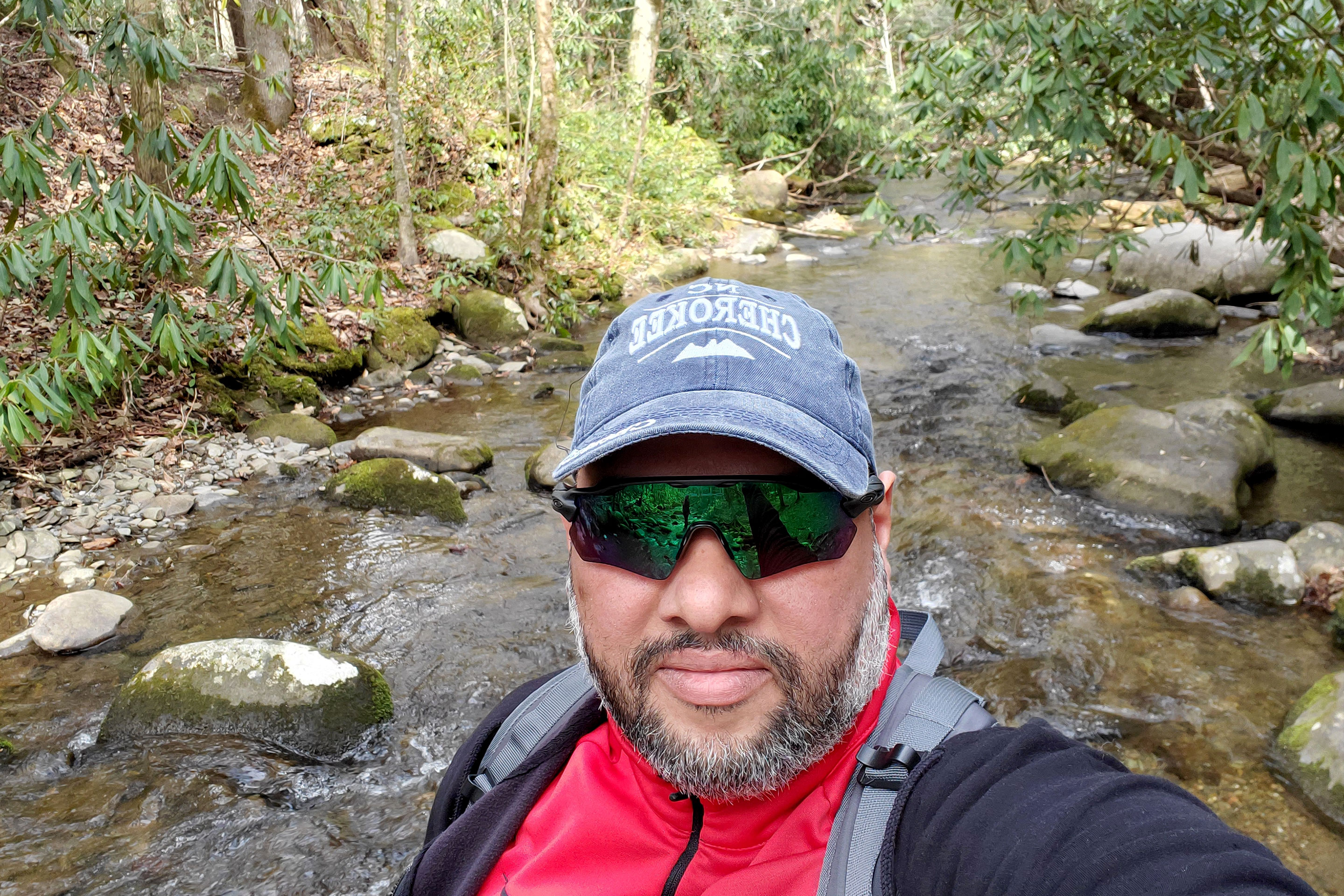 A man wearing a denim baseball cap that reads, 'Cherokee', sunglasses and a navy zip-up hoodie stands in front of a stream in a forest.