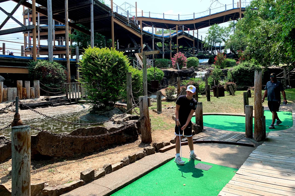 A young boy plays on an outdoor mini golf course surrounded by a wooden roller coaster.