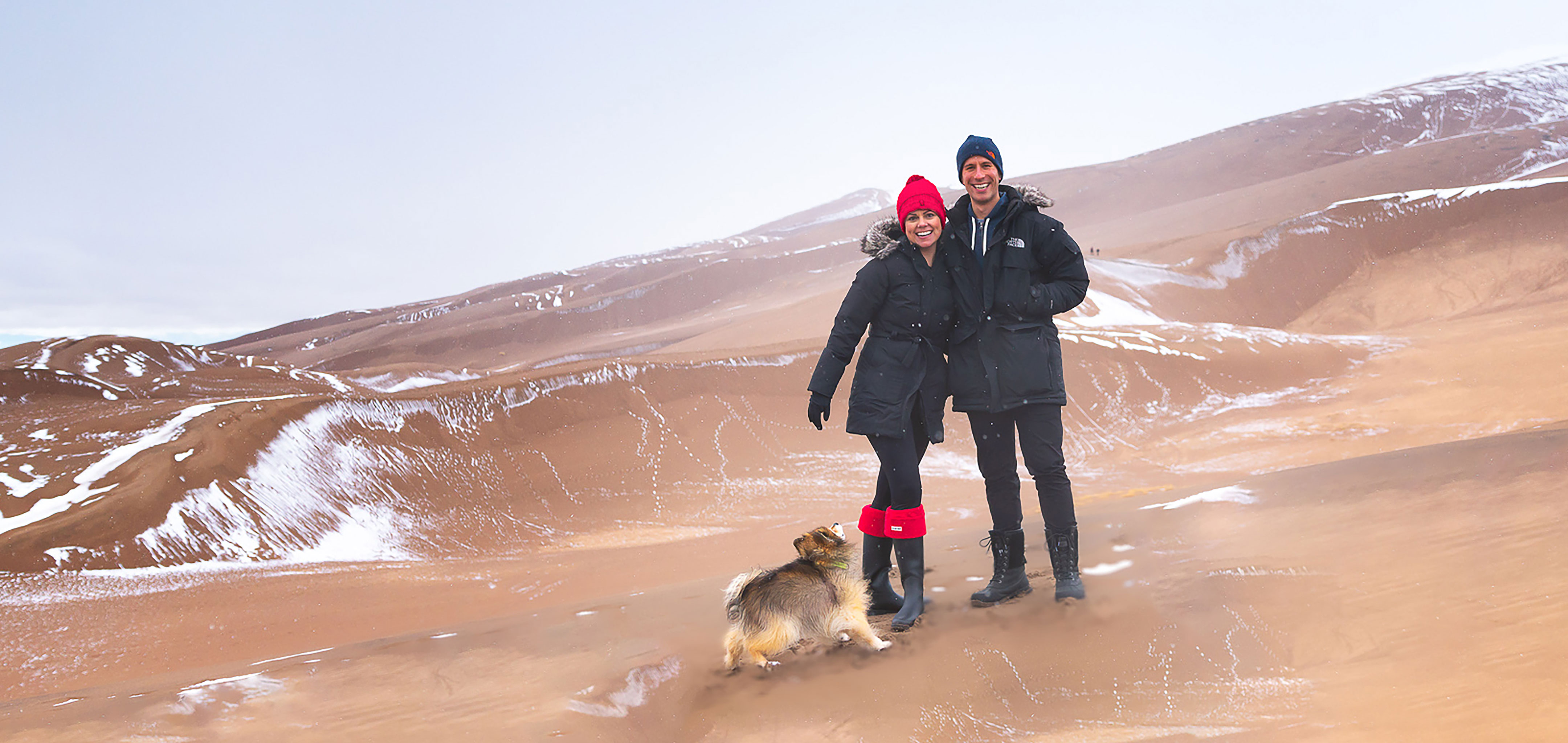 Authors, Lauren Layne and Anthony LeDonne, stand in winter apparel with Bailey the Pomeranian at the Great Sand Dunes National Park and Preserve.