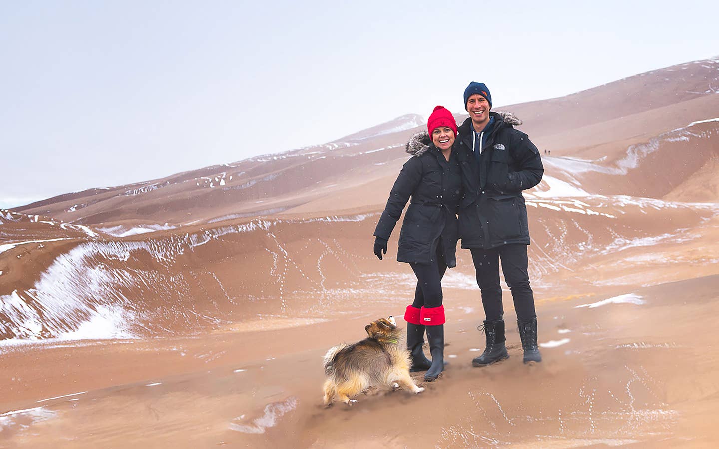 Authors, Lauren Layne and Anthony LeDonne, stand in winter apparel with Bailey the Pomeranian at the Great Sand Dunes National Park and Preserve.