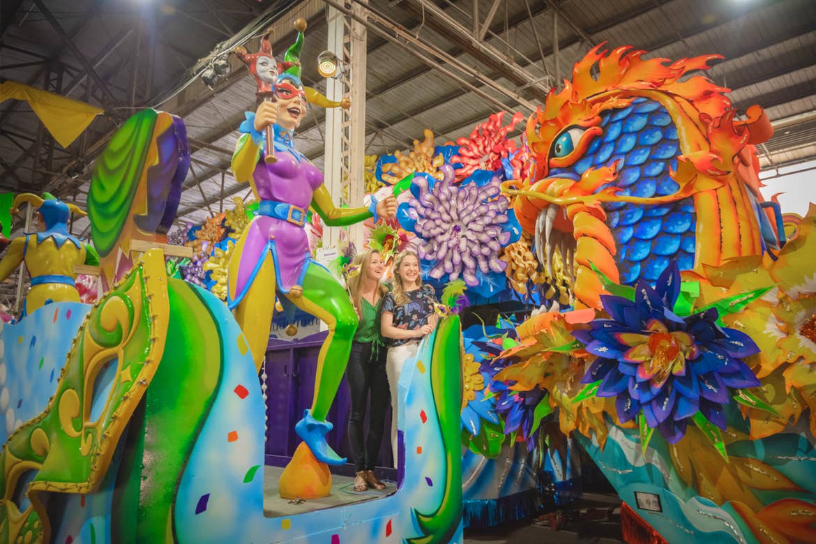 Two caucasian women stand on a retired and vibrant Mardi Gras parade float decorated with dragons and a jester in a warehouse.