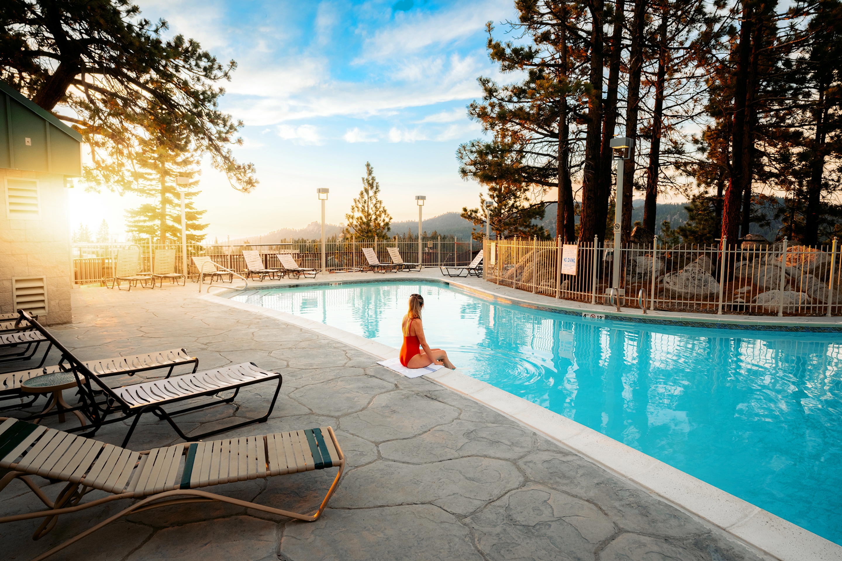 A caucasian woman wears a red one-piece swimsuit as she sits at the edge of a pool dipping her toes in surrounded by sunlight, cloudy blue sky and pine trees.