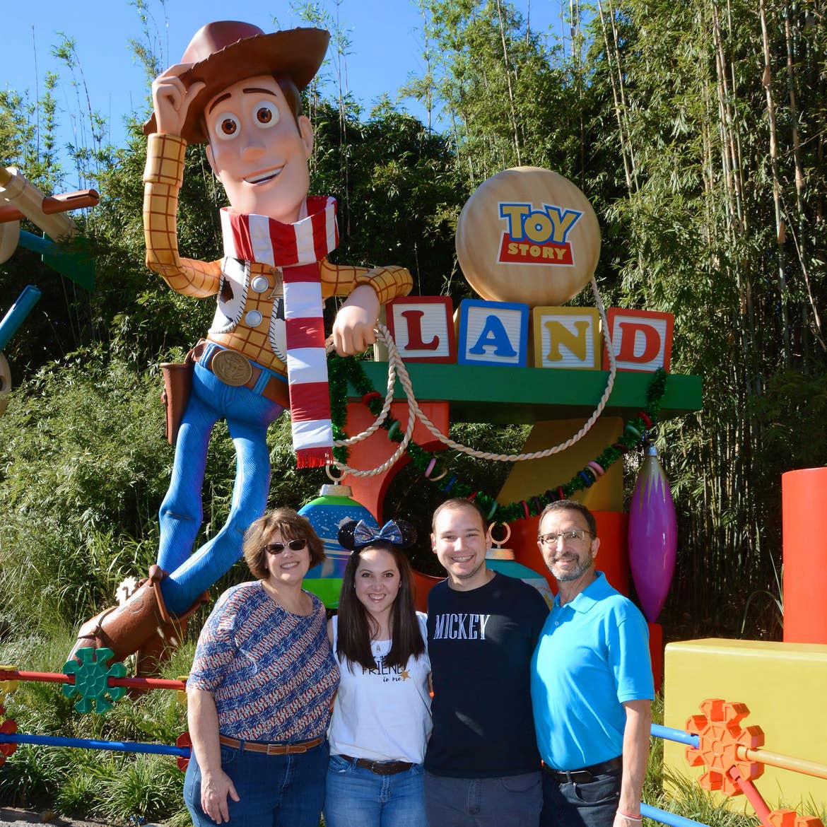 Two women and two men stand next to a scaled version of Sheriff Woody from 'Toy Story' leaning on a sign that reads 'Toy Story Land' made of toys.