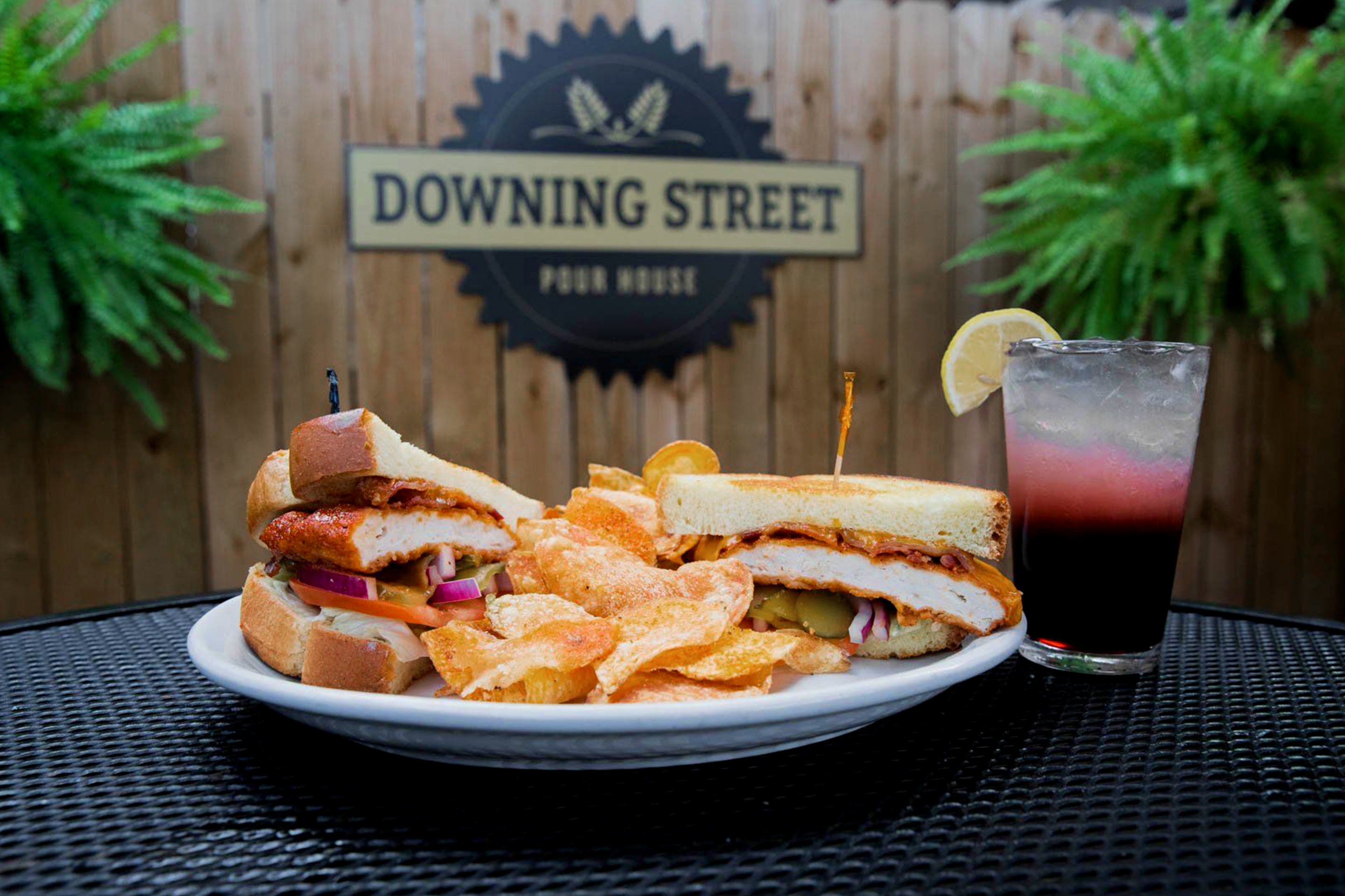 A sandwich split in half, with kettle cooked potato chips are placed on a white plate near a sangria and a picket fence with 'Downing street Pour House' written on it.