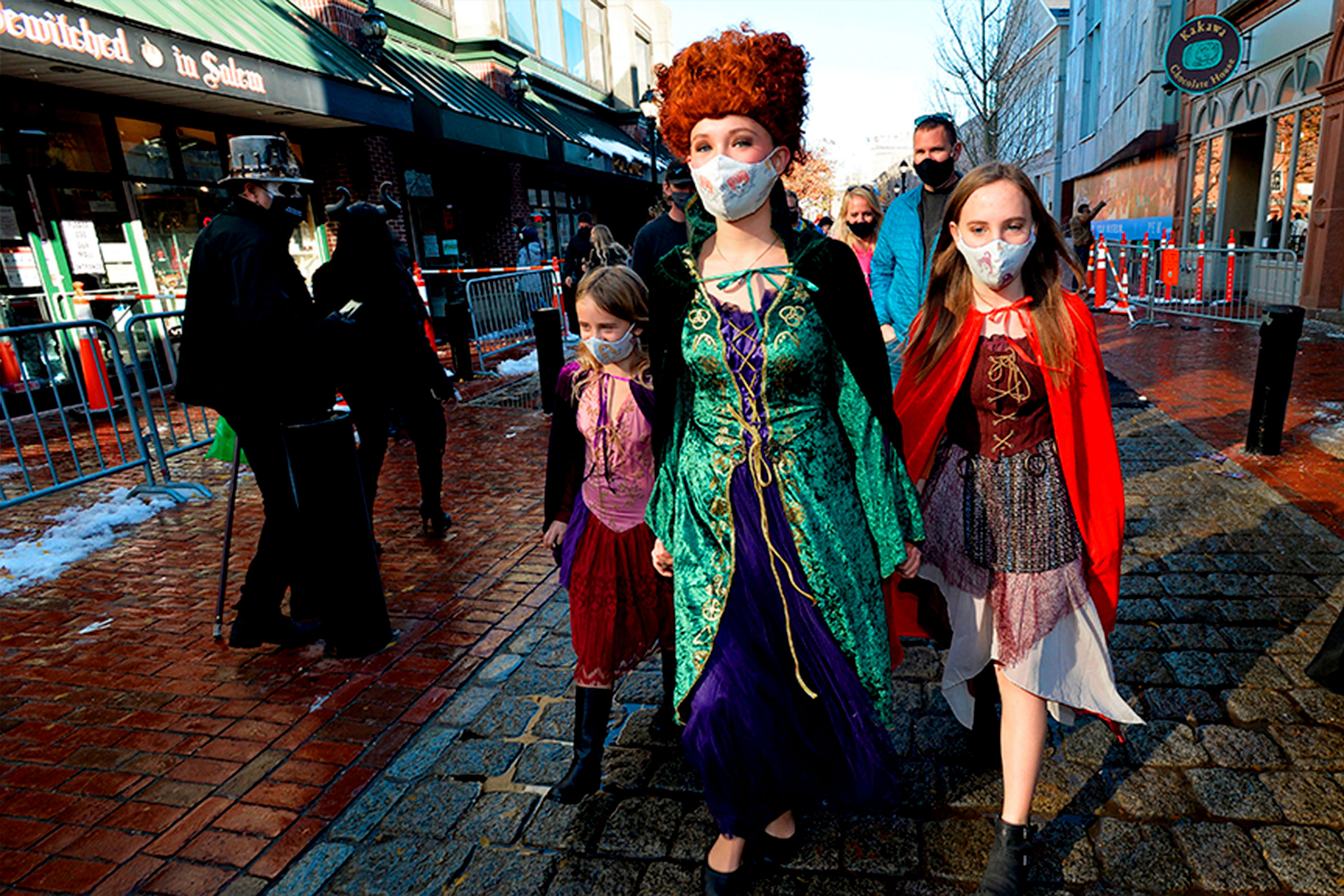 A woman (middle) and two girls (left and right) walk down a brick-paved road wearing safety masks and witch costumes.