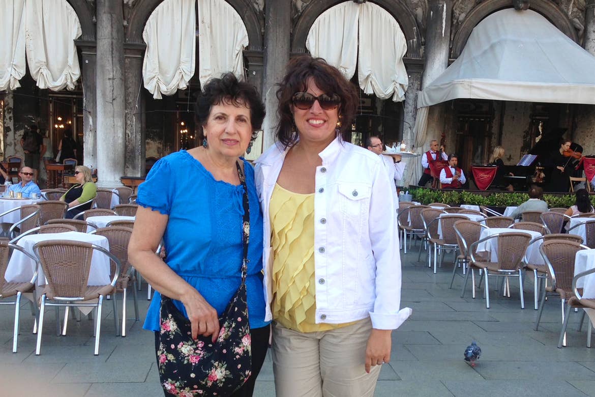 Author, Jennifer Probst (right), wears a white denim jacket and yellow blouse stands next to her mother (left) wearing a blue blouse in front of a restaurant in Venice, Italy.