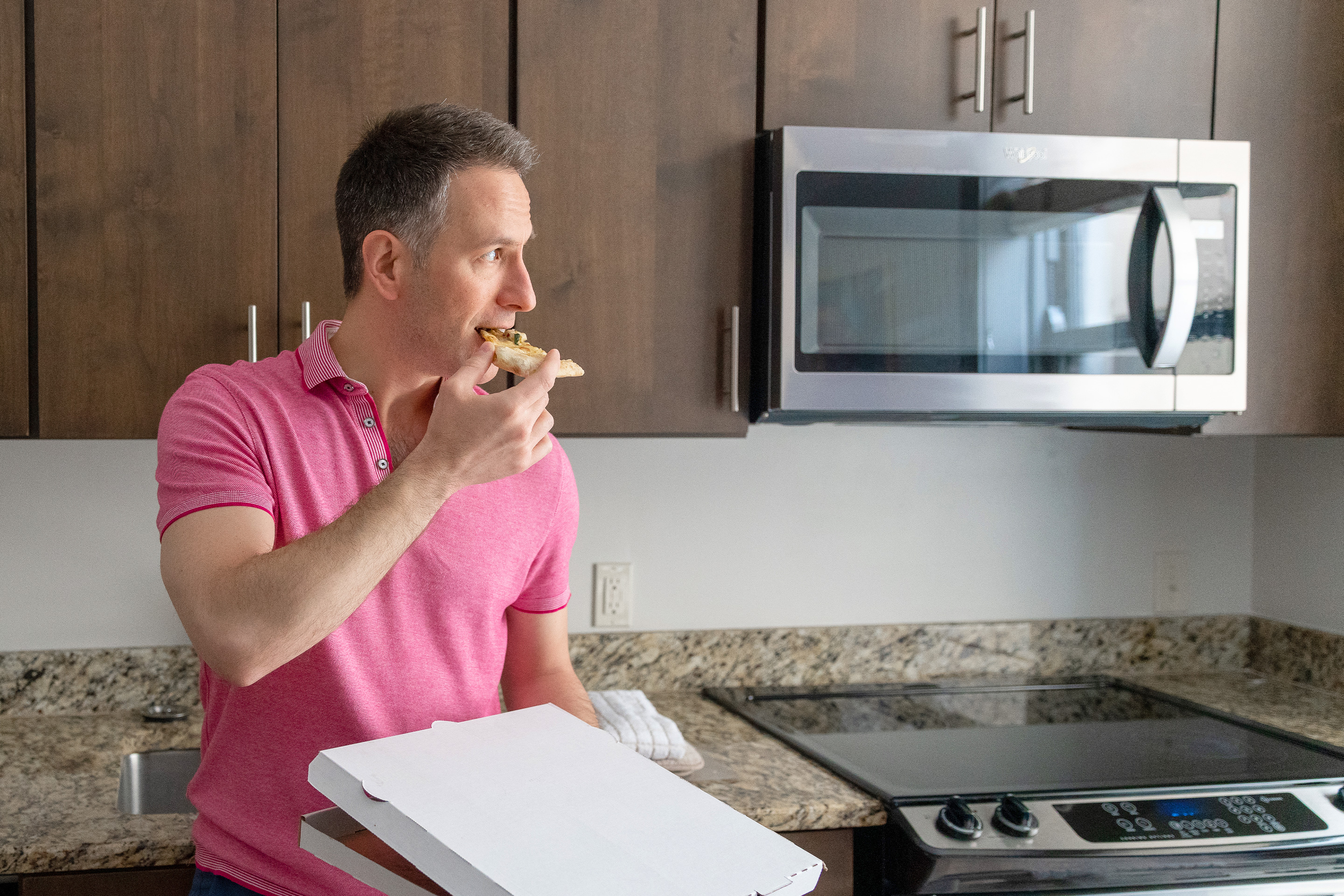 A man (left) wears a pink polo shirt while eating a pizza slice in his right hand and holding a white pizza box in his left hand as he leans on a kitchen countertop.