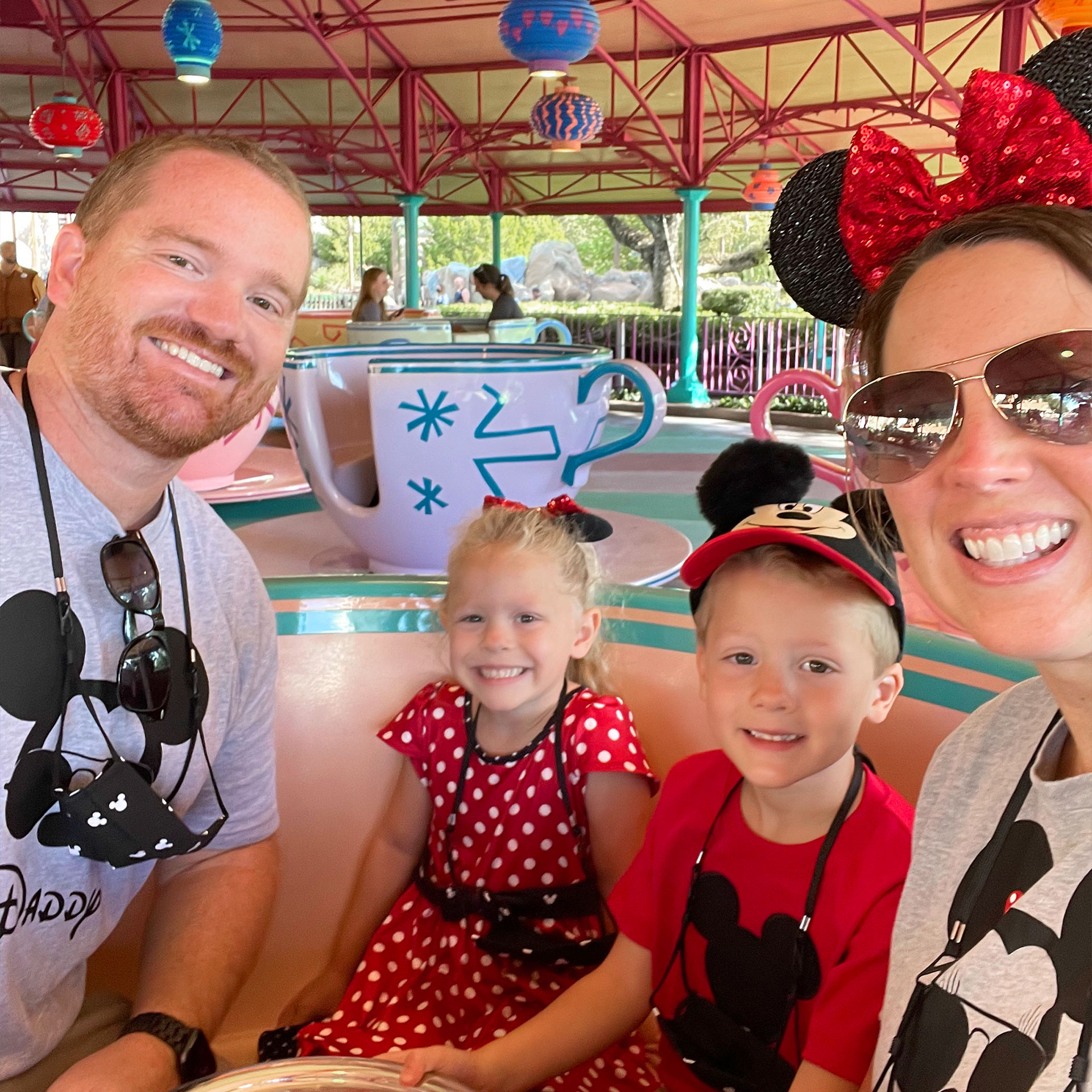 A man, girl, boy and woman sit in a spinning teacup attraction under an enclosed gazebo.