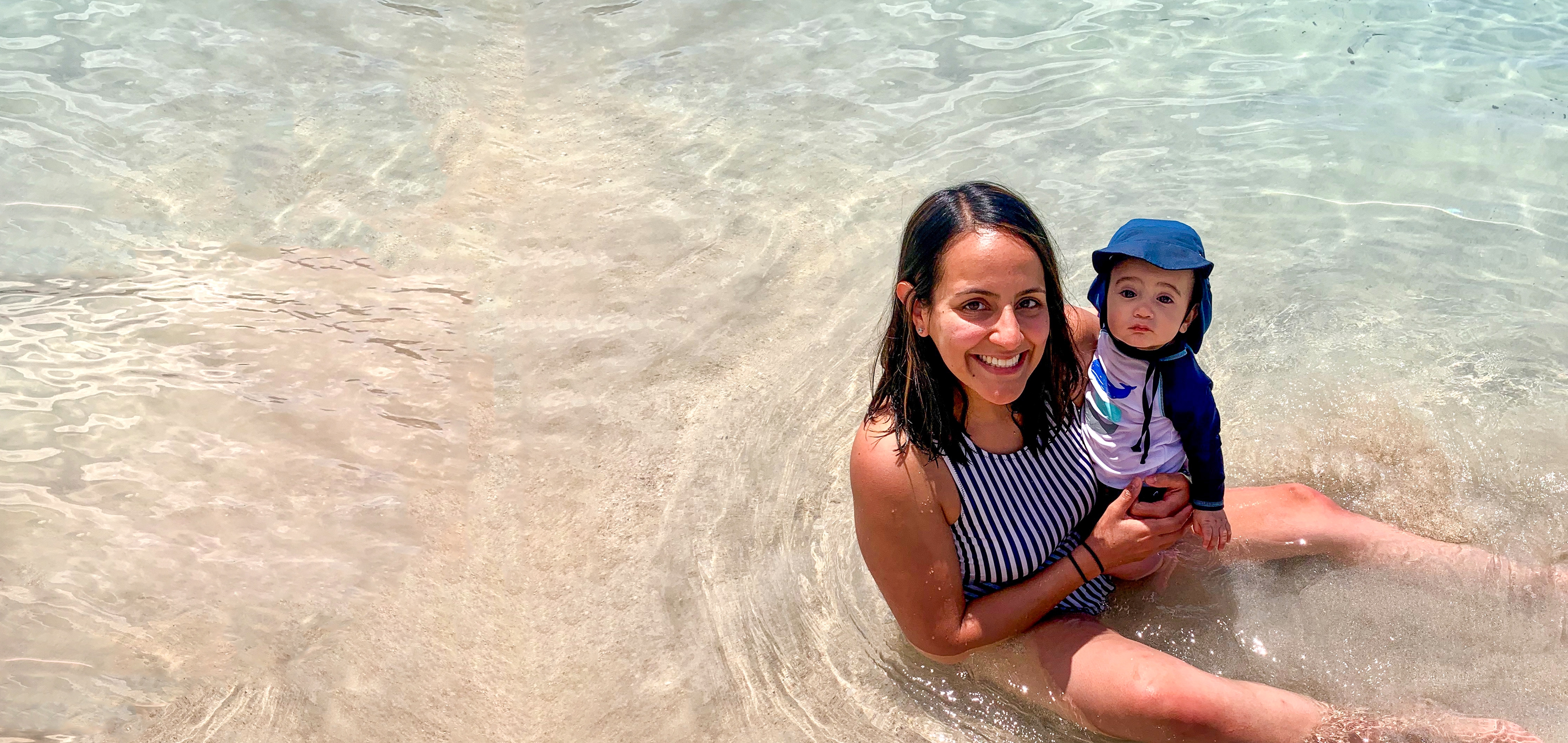 A woman in a striped swimsuit holds a baby wearing swimsuit and a sunhat in shallow water.
