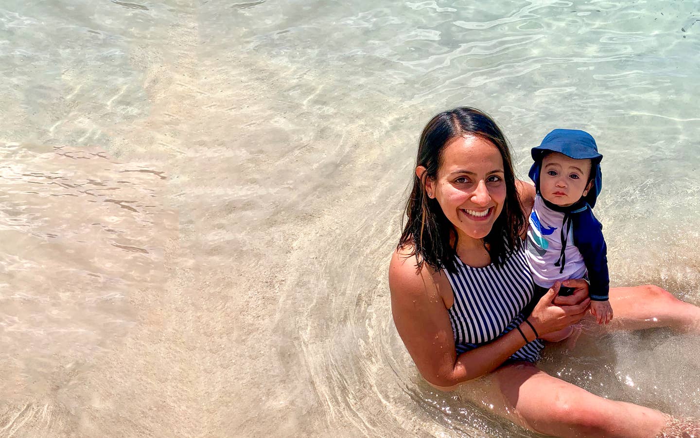 A woman in a striped swimsuit holds a baby wearing swimsuit and a sunhat in shallow water.