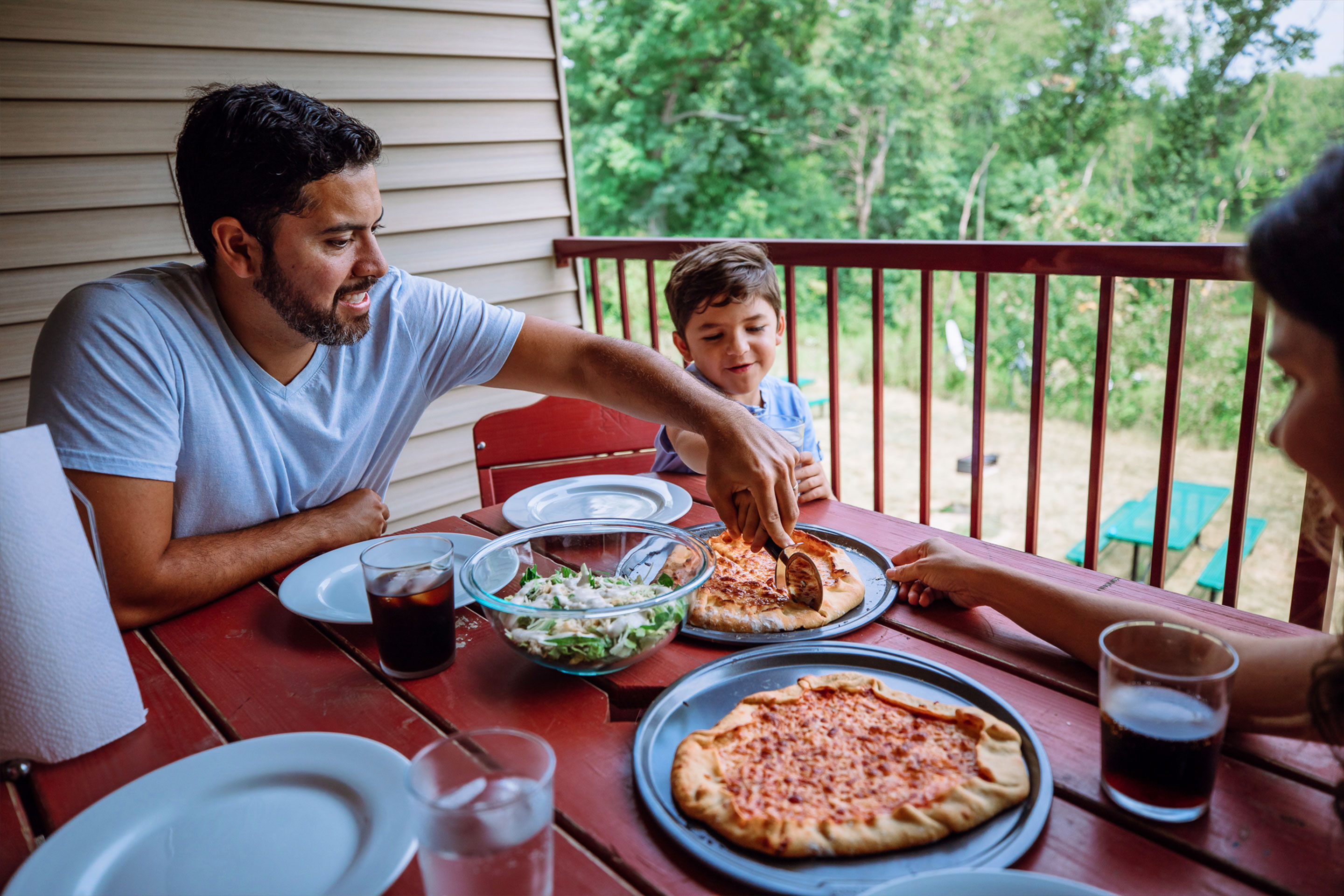 From left to right: A man cuts slices of pizza as a young boy and woman sit on an outdoor patio at a red, wood table.