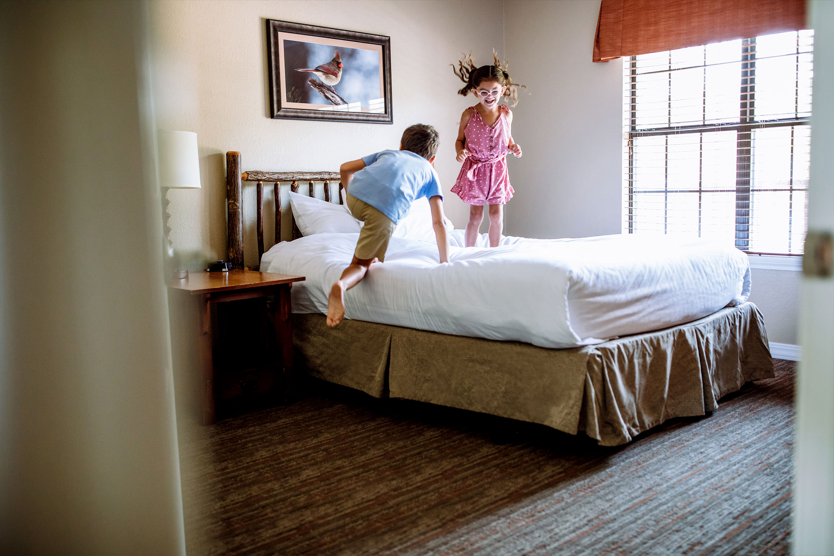 A young boy (left) and young girl (right) jump on the bed in a villa.
