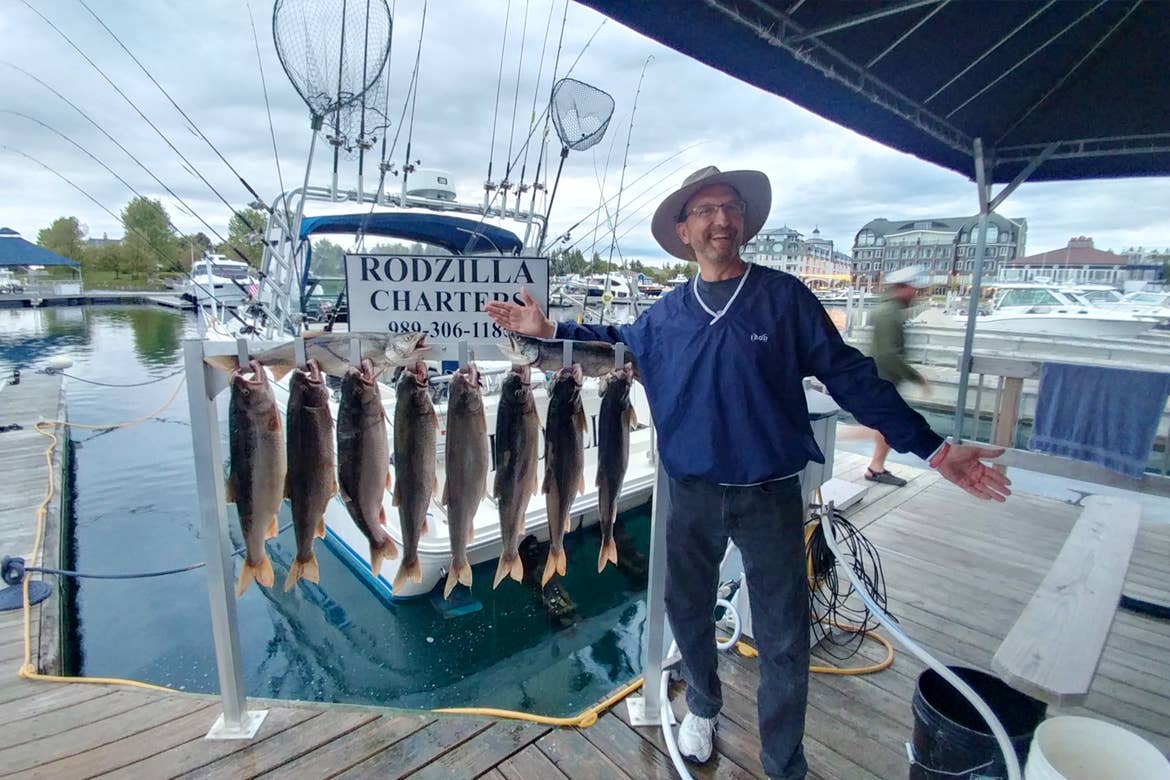 A caucasian man wearing a sun hat, navy windbreaker and jeans stands on a dock near some freshly caught fish with a white sign that reads, 'Rodzilla Charters.'