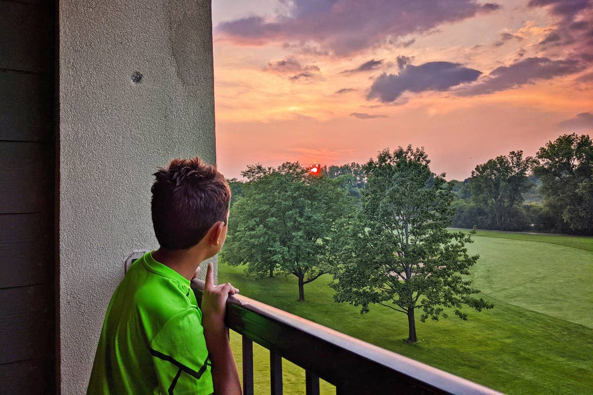 A young boy stands on a balcony overlooking a sunset.