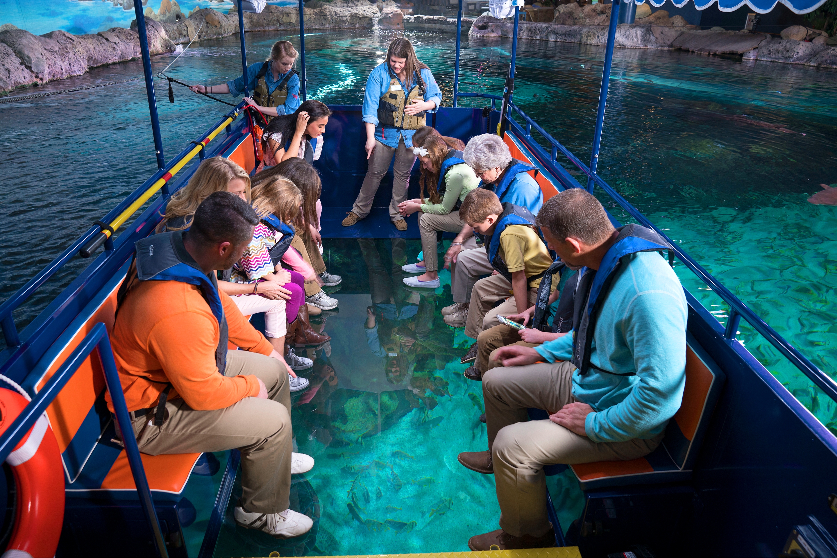 Guests sit on a glass-bottom boat wearing life jackets at an aquarium.