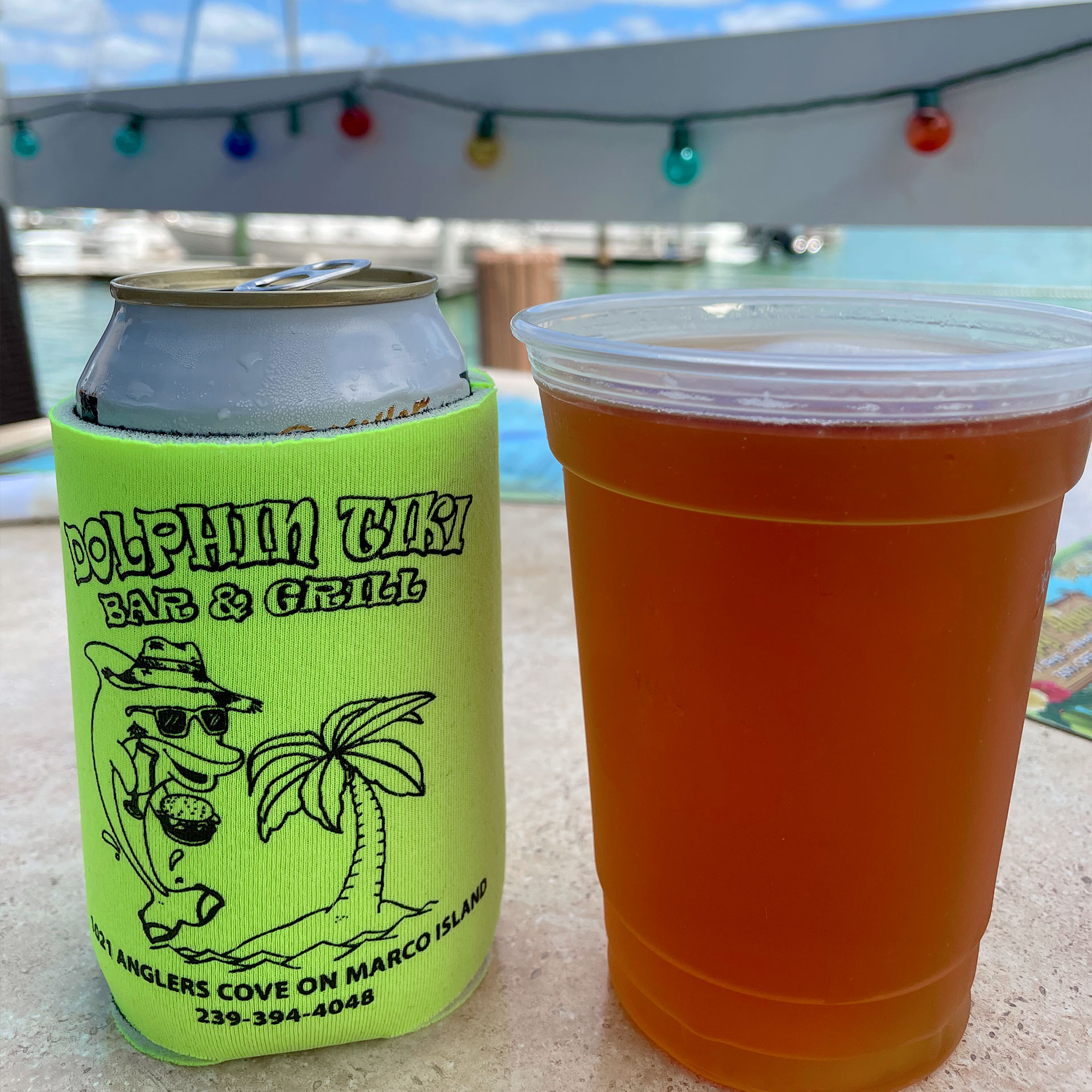 Two alcoholic beverages, one in an aluminum can with themed coozy and the other in a plastic cup, sit on a table outdoors near a white fence with Christmas string lights.