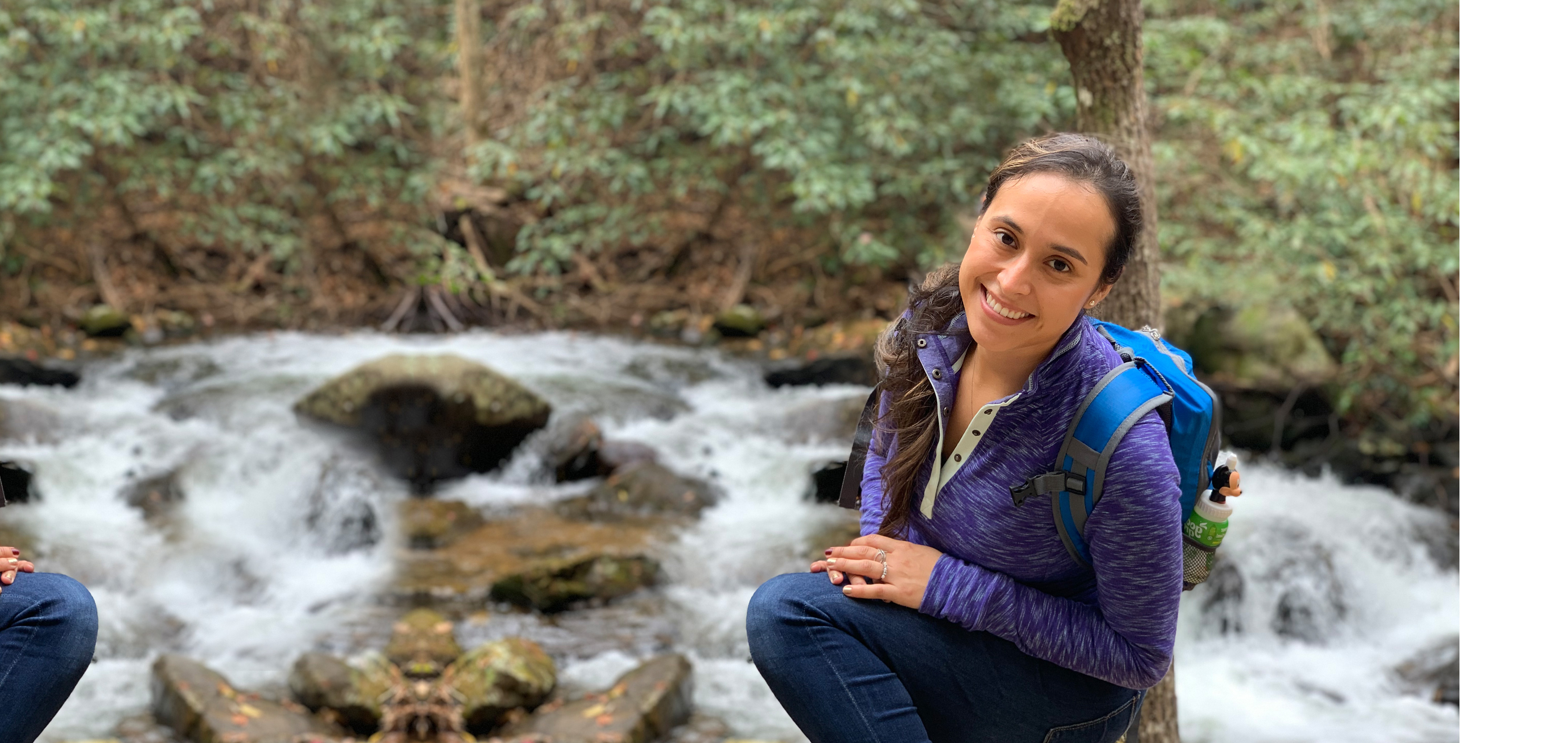 Featured author, Andrea Beltran, poses near a creek wearing a blue hiking backpack and purple pull-up sweater.