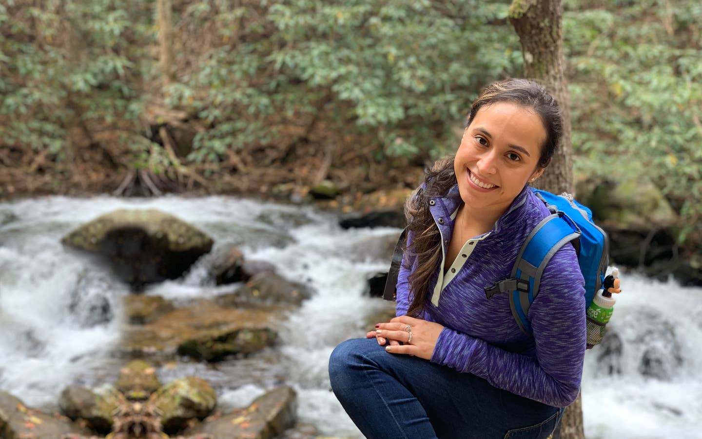 Featured author, Andrea Beltran, poses near a creek wearing a blue hiking backpack and purple pull-up sweater.