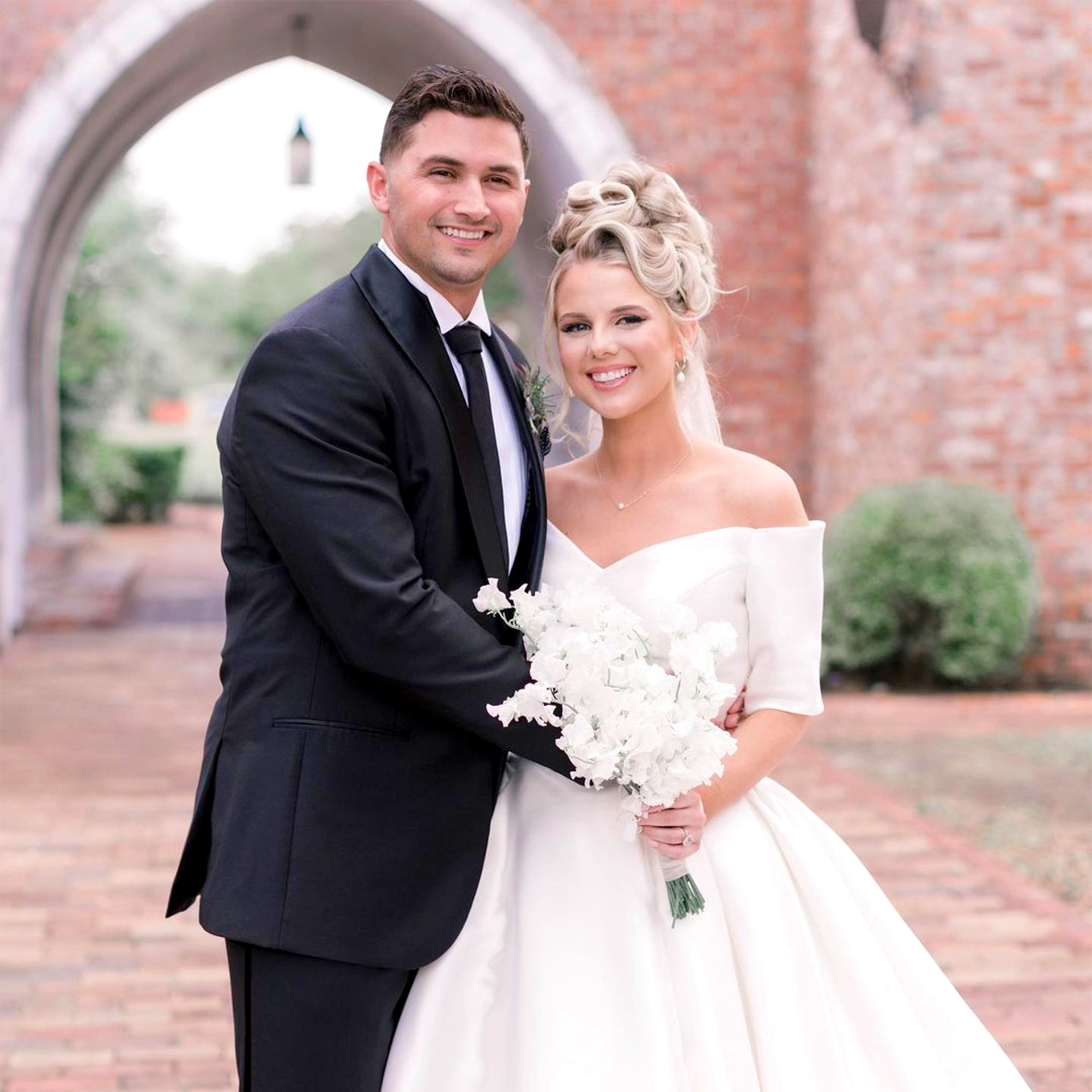 A Latinx groom wearing a black suit and necktie (left) and a caucasian bride with an updo (right) pose next to each other in front of a brick facade.
