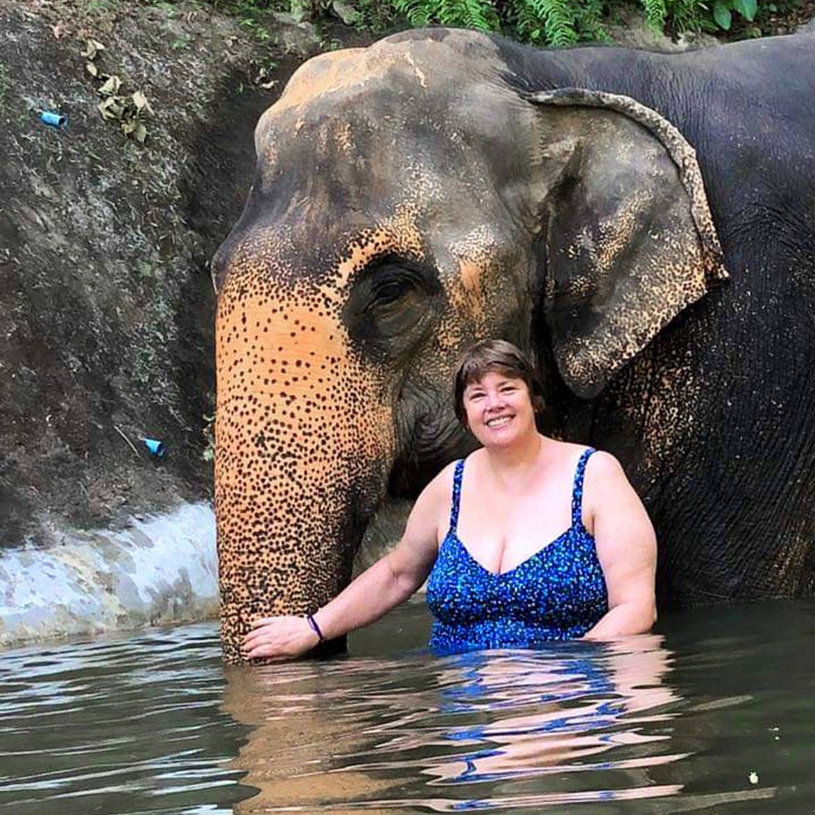 A caucasian woman wearing a blue swimsuit stands near an elephant submerged in water in Phuket, Thailand.