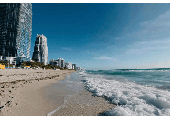 Waves splash white onto shoreline with tall hotel buildings in the distance.