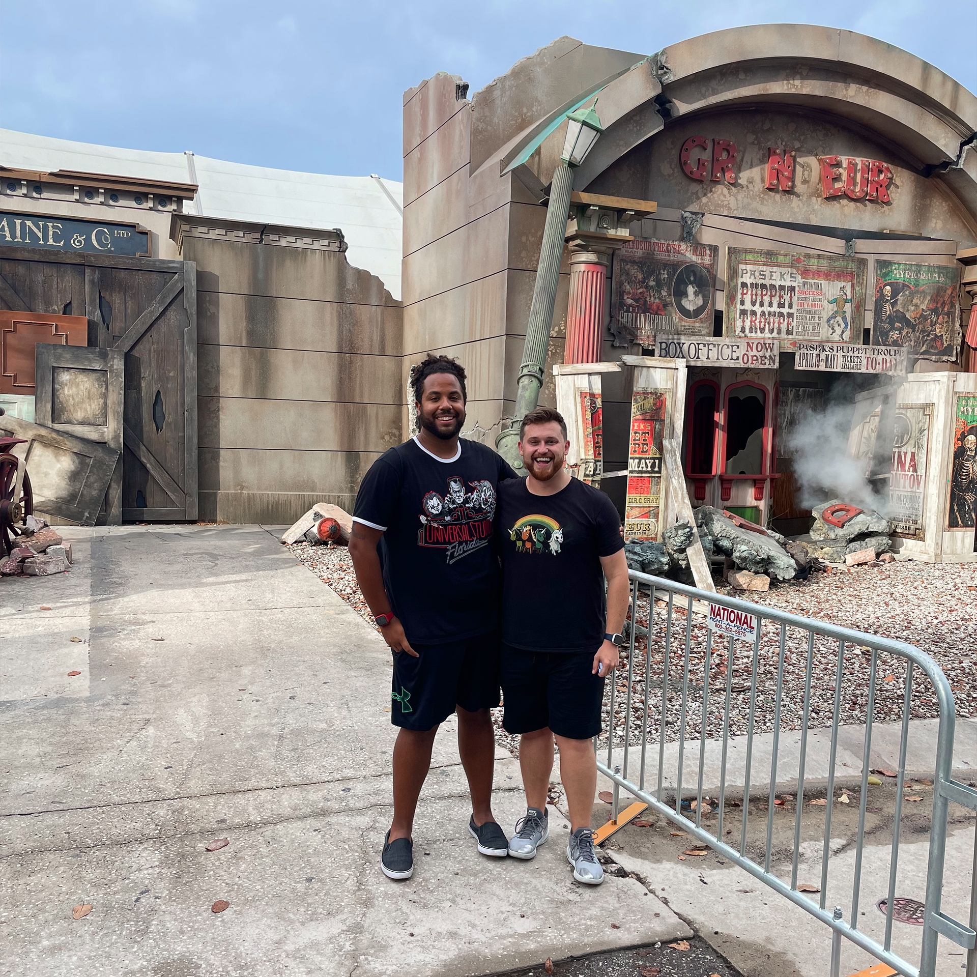Two men wearing black t-shirts and shorts stand in front of a haunted house.