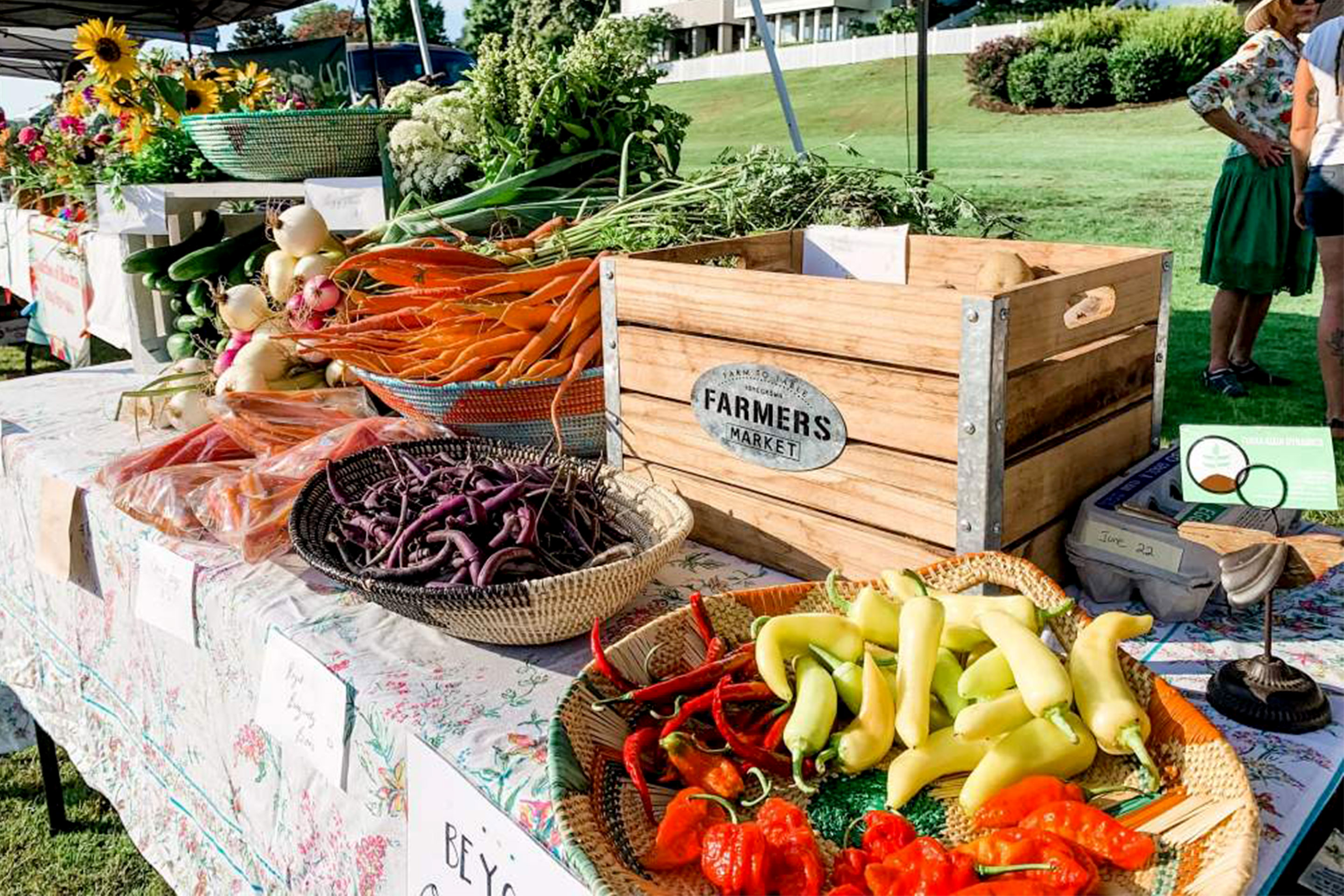 A wooden crate basket is surrounded by various produce at an outdoor farmers market.