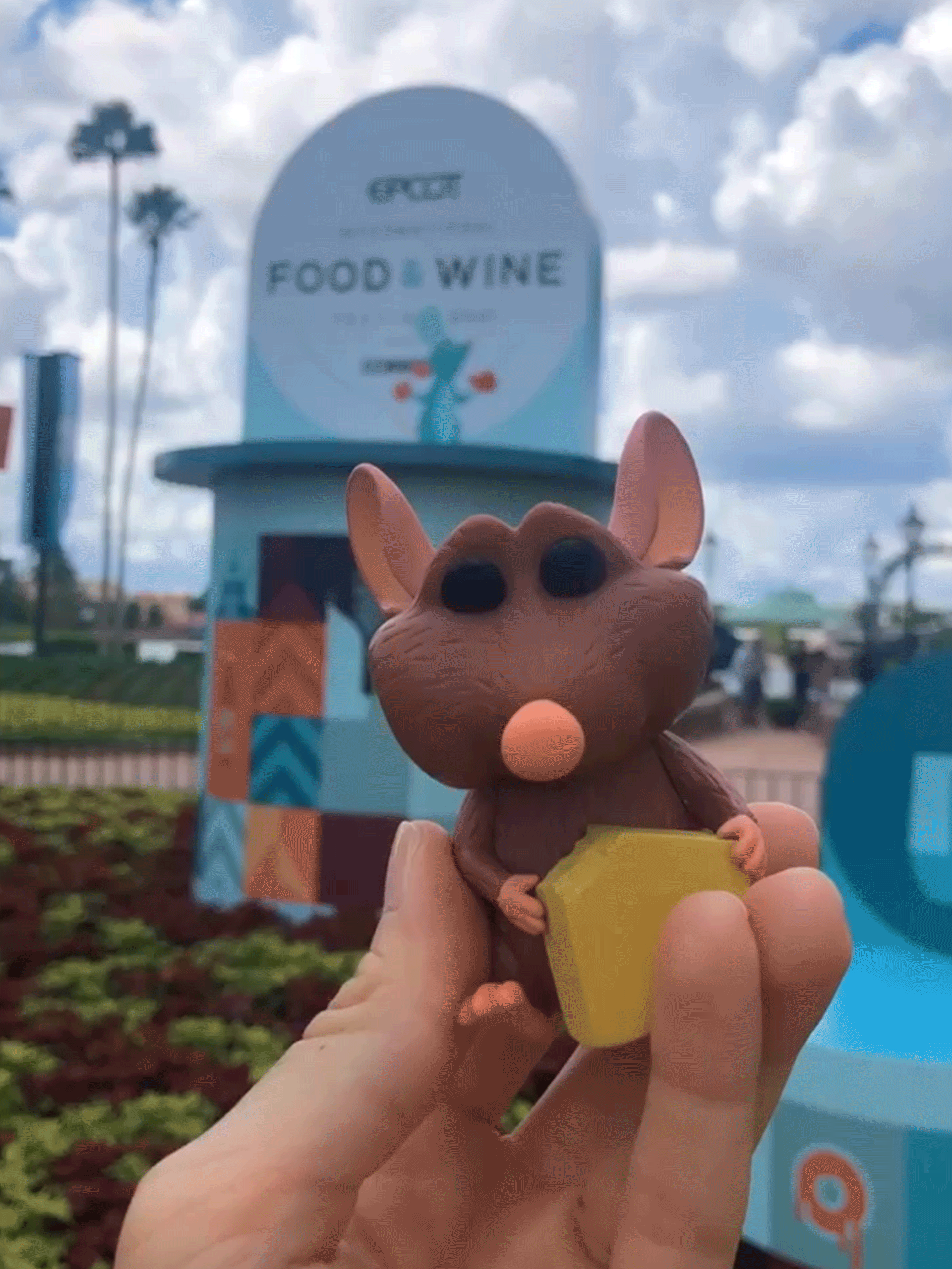 A hand holds an Emile Funko pop figurine near a garden sign display that reads, 'EPCOT Food & Wine Festival.'