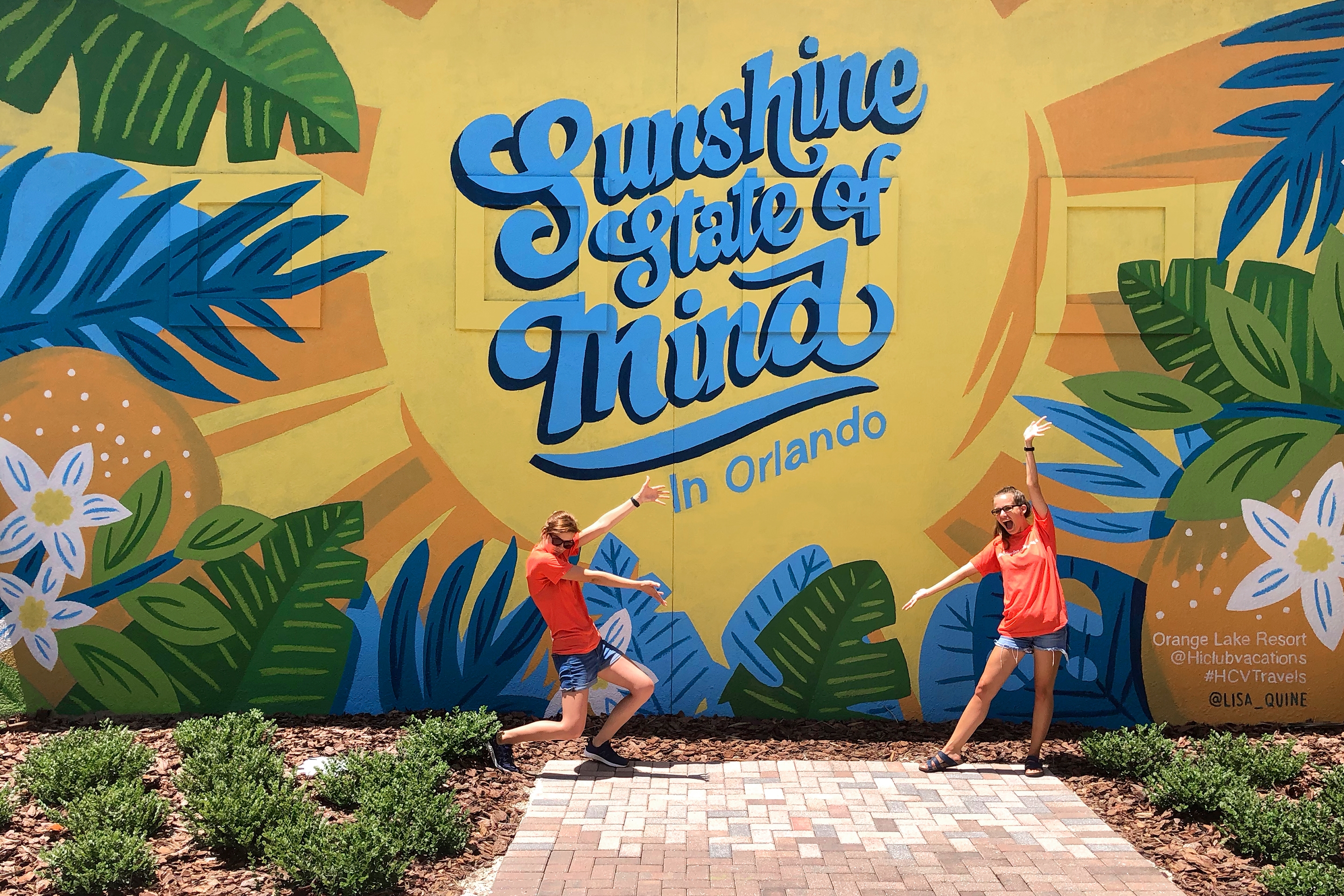 Two caucasian women wearing orange t-shirts and denim shorts stand with their arms extended at our mural located in North Village at Orange Lake Resort in Orlando, Florida.