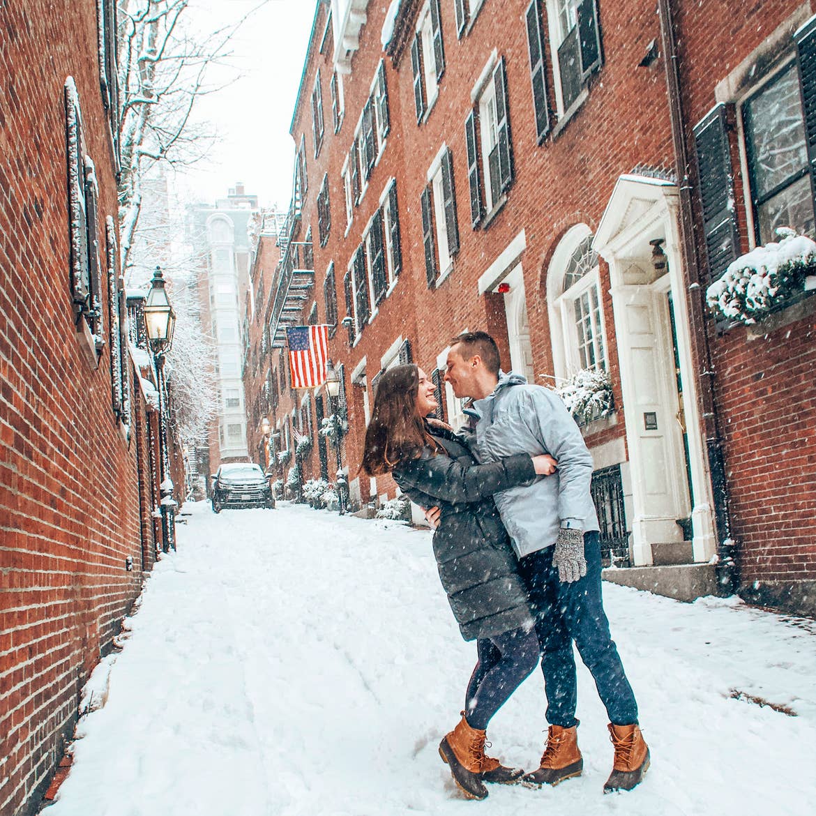 A woman in a green puffer jacket and a man in a white-grey jacket embrace on a snow-covered street near historical brick buildings.