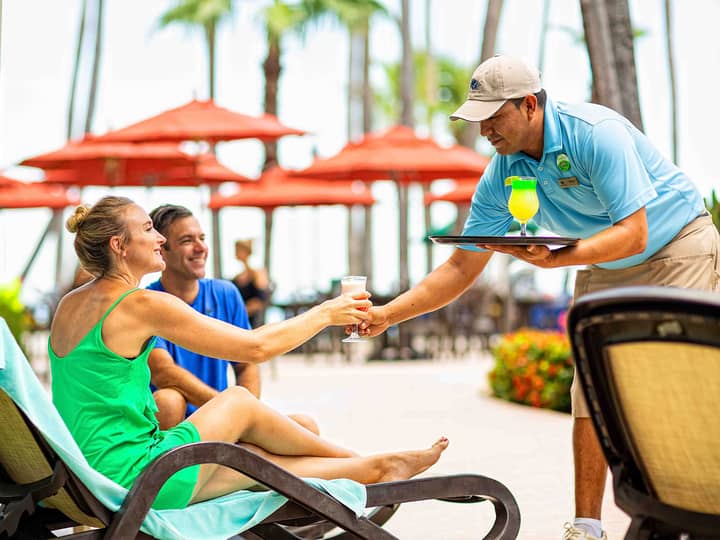 A man handing a cocktail to a woman sitting in a pool chair outside at The Royal Cancun in Cancun, Mexico.