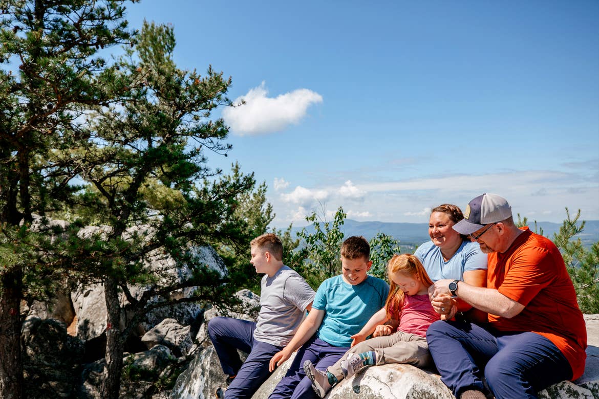 A caucasian family of five (left to right: Two tween boys, a young girl, a woman and man) sit on a rock formation near pine trees at the peak of Monument Mountain in Lee, Massachusetts.