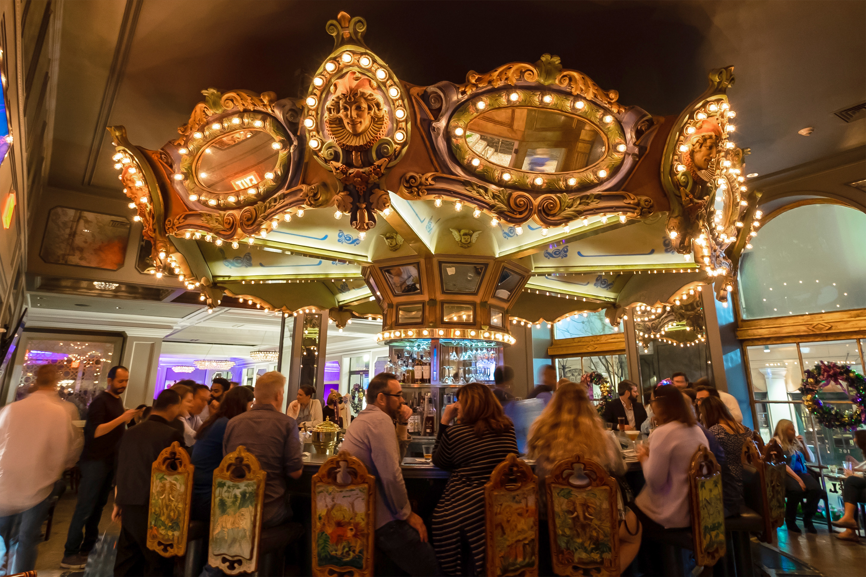 A carousel topper illuminated with rounded light bulbs is anchored above a rounded bar surrounded by patrons.