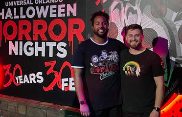 Two men wearing black t-shirts and shorts stand in front of a mural at night.