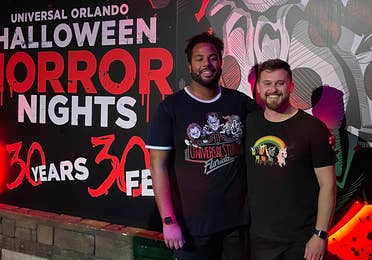 Two men wearing black t-shirts and shorts stand in front of a mural at night.
