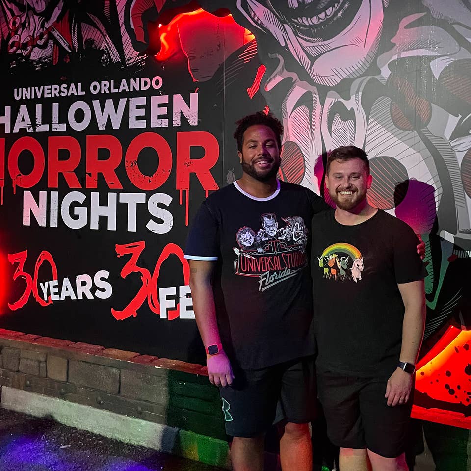 Two men wearing black t-shirts and shorts stand in front of a mural at night.