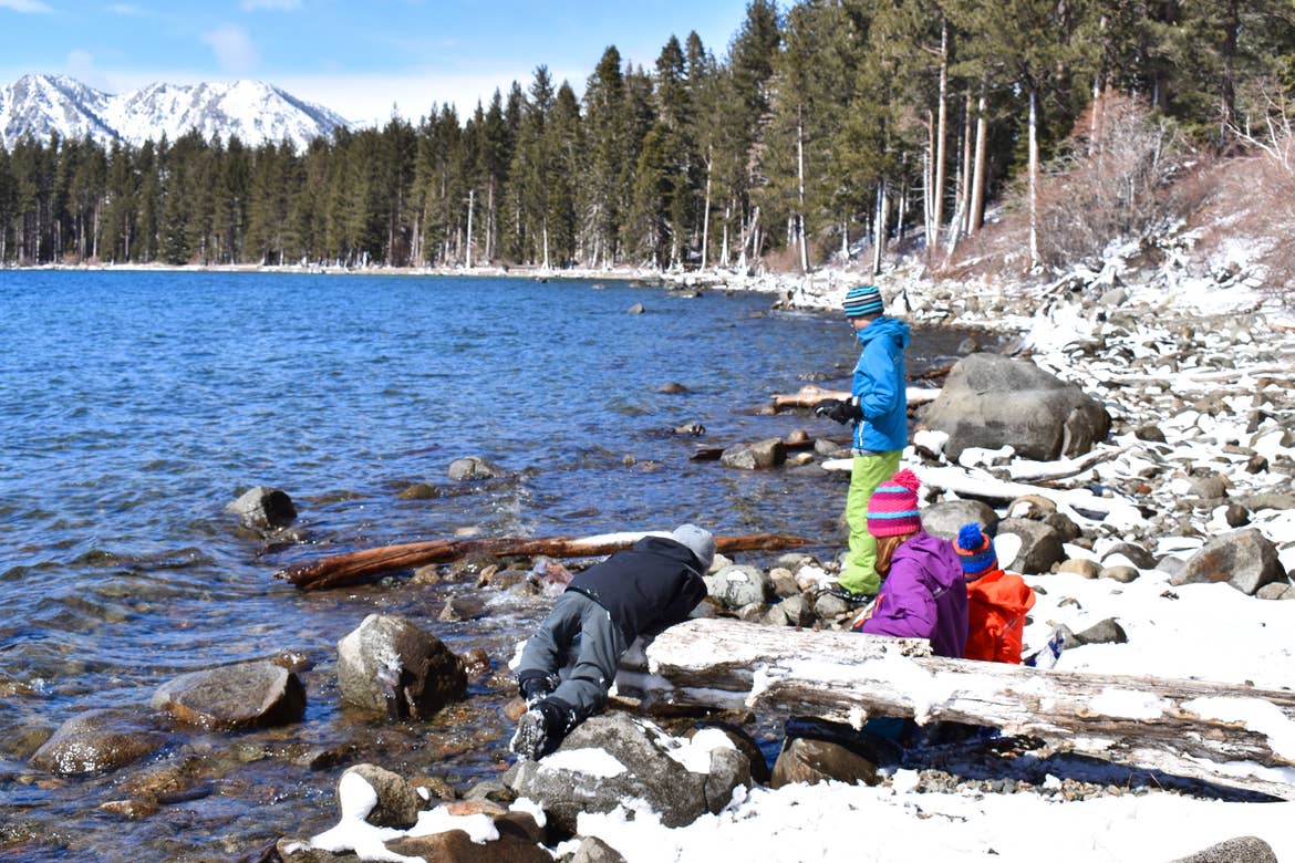 Four children stand near a lake wearing winter apparel and throwing rocks.