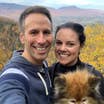 Authors, Lauren Layne and Anthony LeDonne, stand in front of fall foliage with Bailey the Pomeranian.
