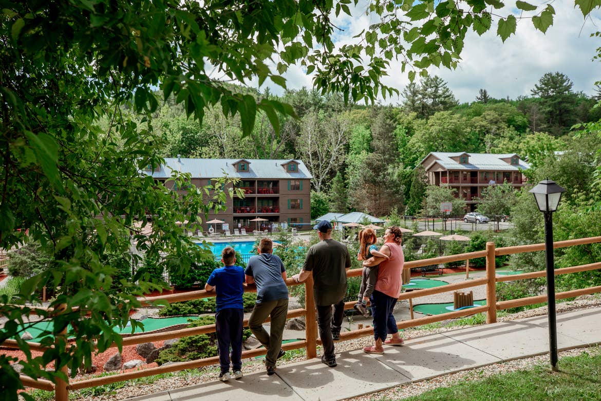 A caucasian family of five (Left to Right: Two tween boys, a man, a woman holding a young girl) stand near a fence that overlooks our Oak and Spruce resort mini-golf course, main pool, basketball courts and villa exteriors.