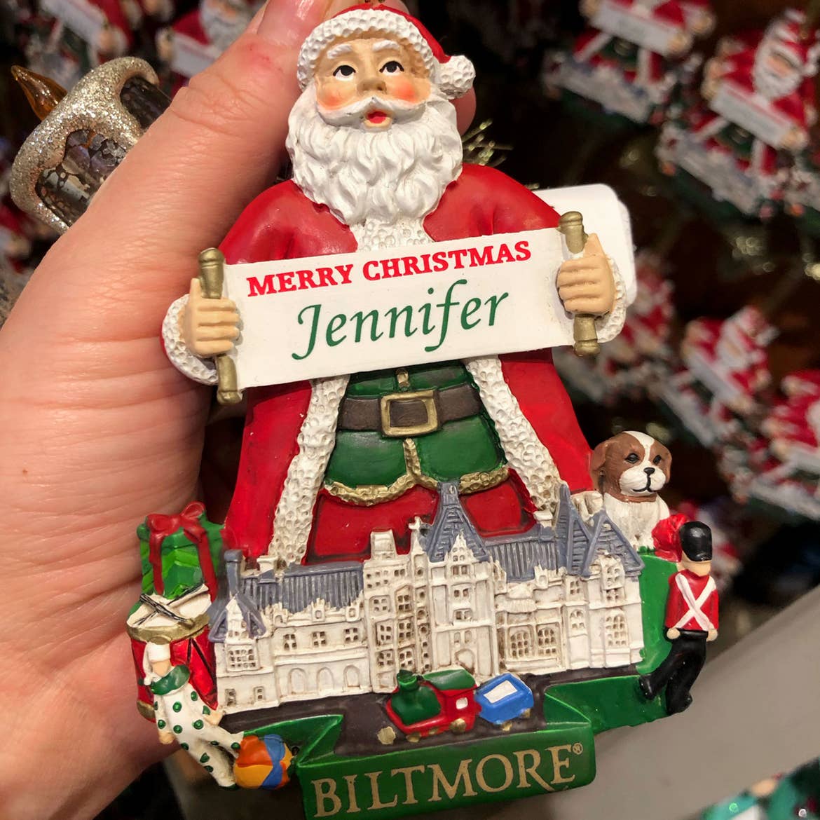 Author, Jenn C. Harmon, holds a Santa Claus ornament which holds a sign that reads, 'Merry Christmas Jennifer' standing behind the silhouette of the Biltmore Estate.