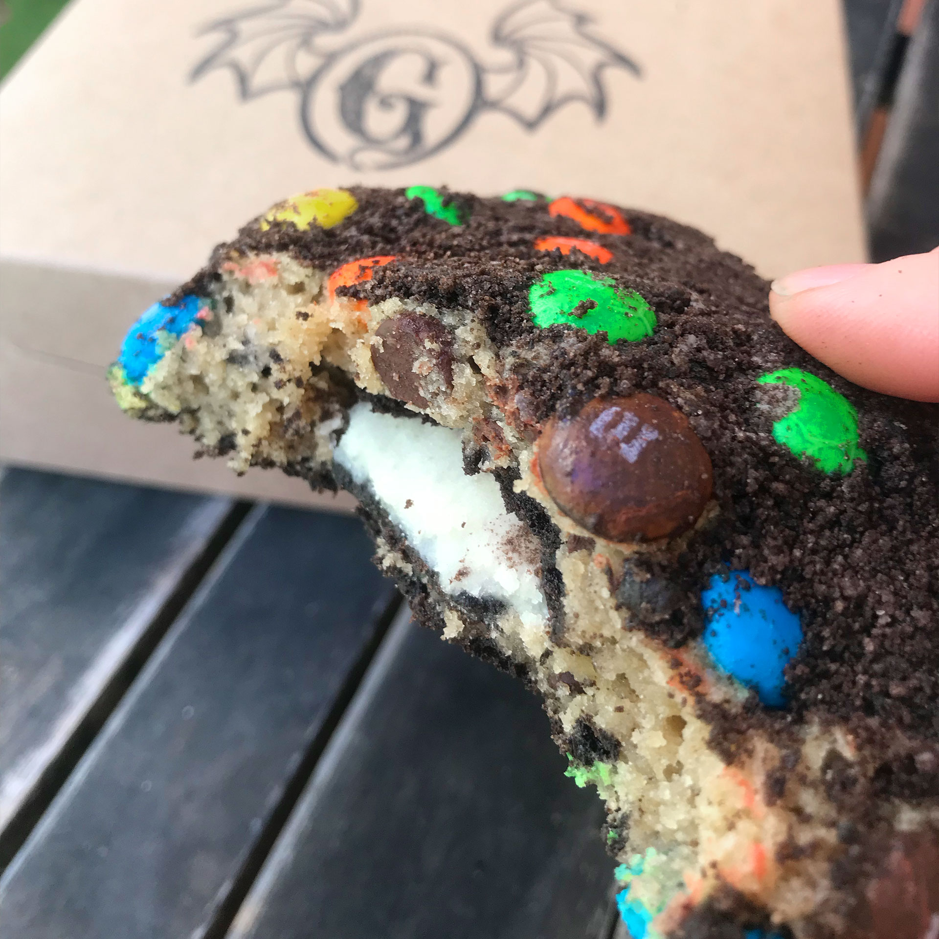 An m&m encrusted chocolate chip cookie with an Oreo cookie baked inside is held by a woman's hand over a brown takeout box.