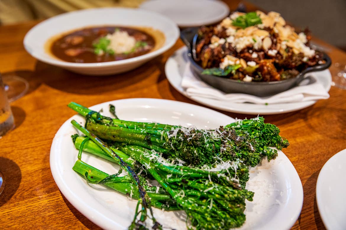 Three white serving dishes containing Seafood Gumbo à La Creole, Crispy Brussels Sprouts, and Charred Broccolini on a wooden dining table.