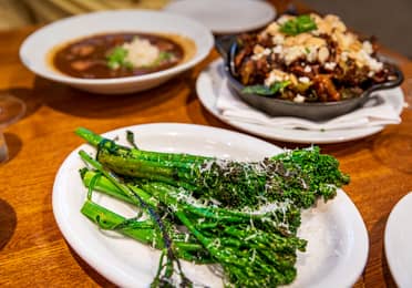 Three white serving dishes containing Seafood Gumbo à La Creole, Crispy Brussels Sprouts, and Charred Broccolini on a wooden dining table.