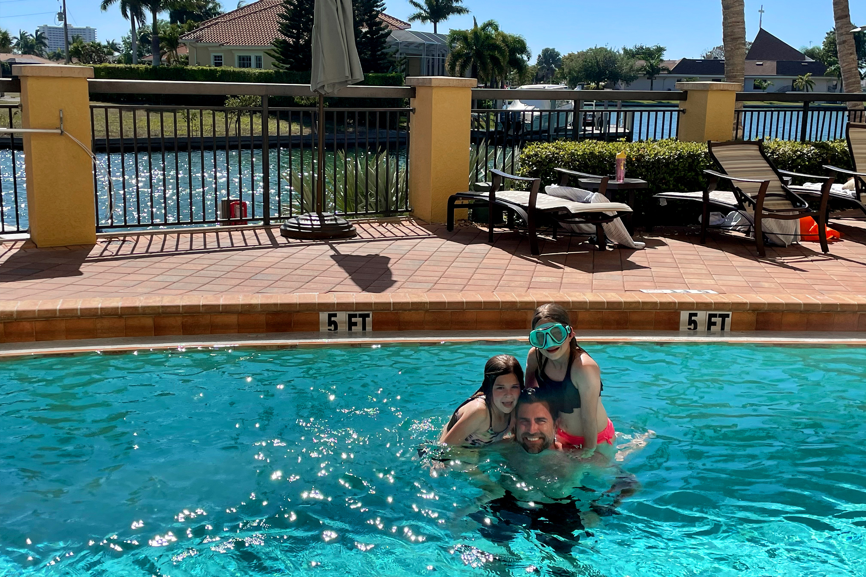Two young caucasian girls (top-left and top-right) wearing swimsuits crouch on the shoulders of a caucasian male (middle) at the outdoor pool entrance at Sunset Cove Resort in san Marco, Florida.