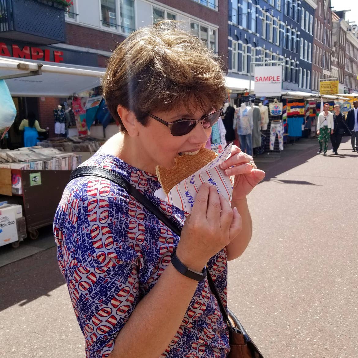 A caucasian woman wearing a patriotic-patterned shirt eats a stroopwaffle in the walkway of the Albert de Cuyp Market in Amsterdam, Netherlands.