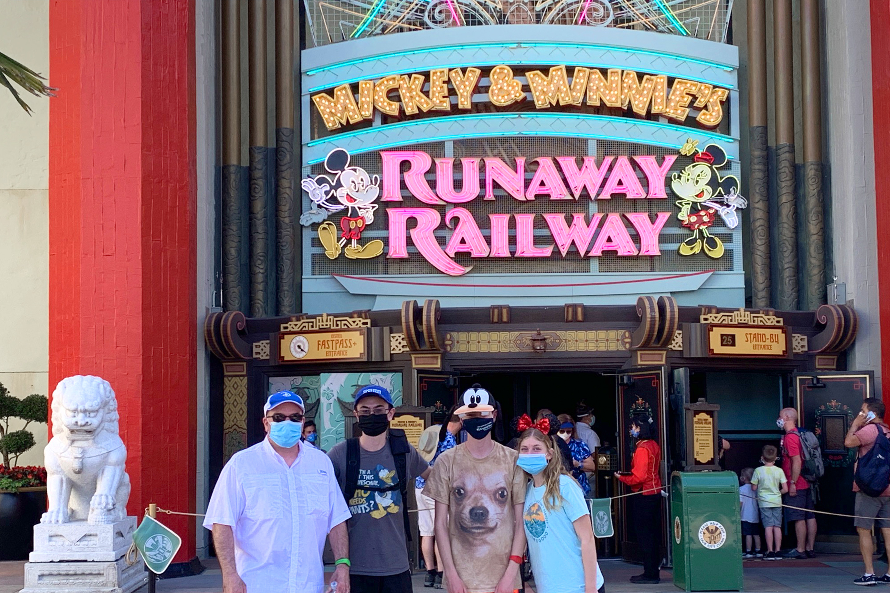 Three men (left) and a young woman (right) stand wearing safety masks outside the ride queue of Mickey & Minnie's Runaway Railway attraction in Disney's Hollywood Studios at Walt Disney World Resort.